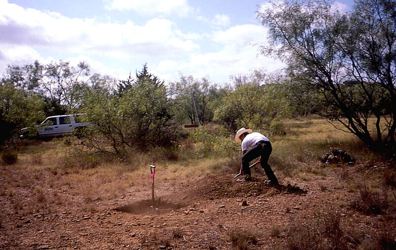 Photo of young man shoveling dirt back into a square hole.