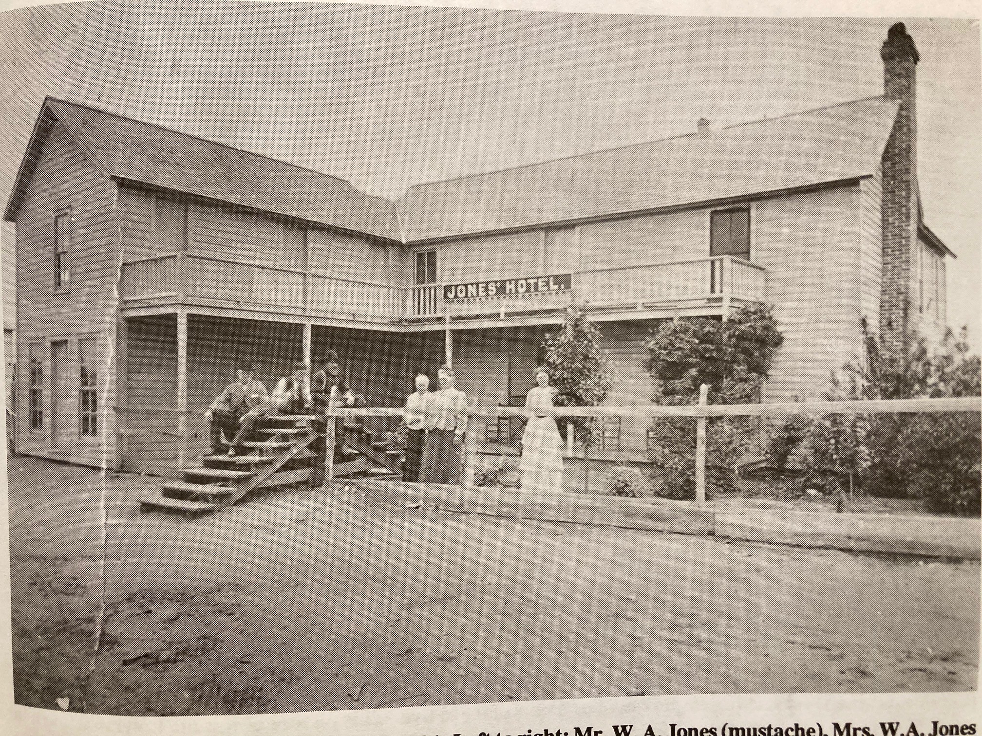 sepia photo of a two-story L-shaped wooden structure with people posing in front of it.