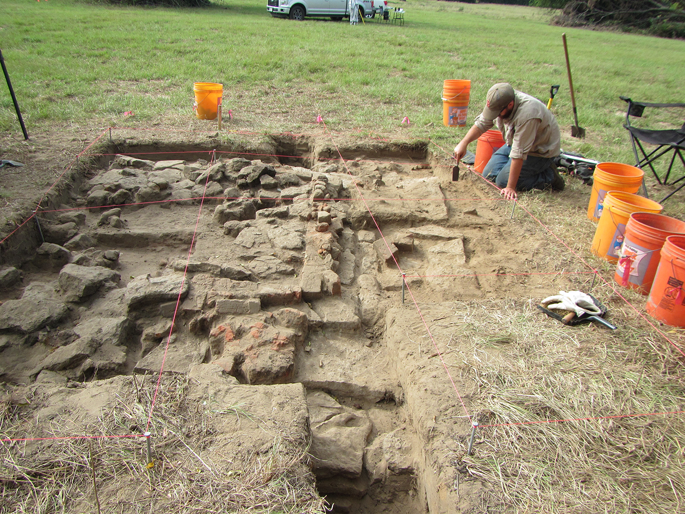 A man kneeling on grass and excavating with a trowel. Several orange buckets are around him. The excavation is shallow, square, and filled with rocks.