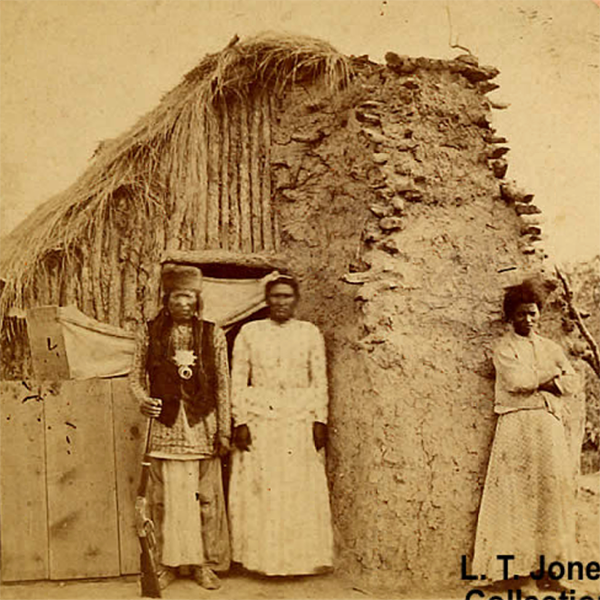 black and white photograph of Native Americans in front of a jacal structure