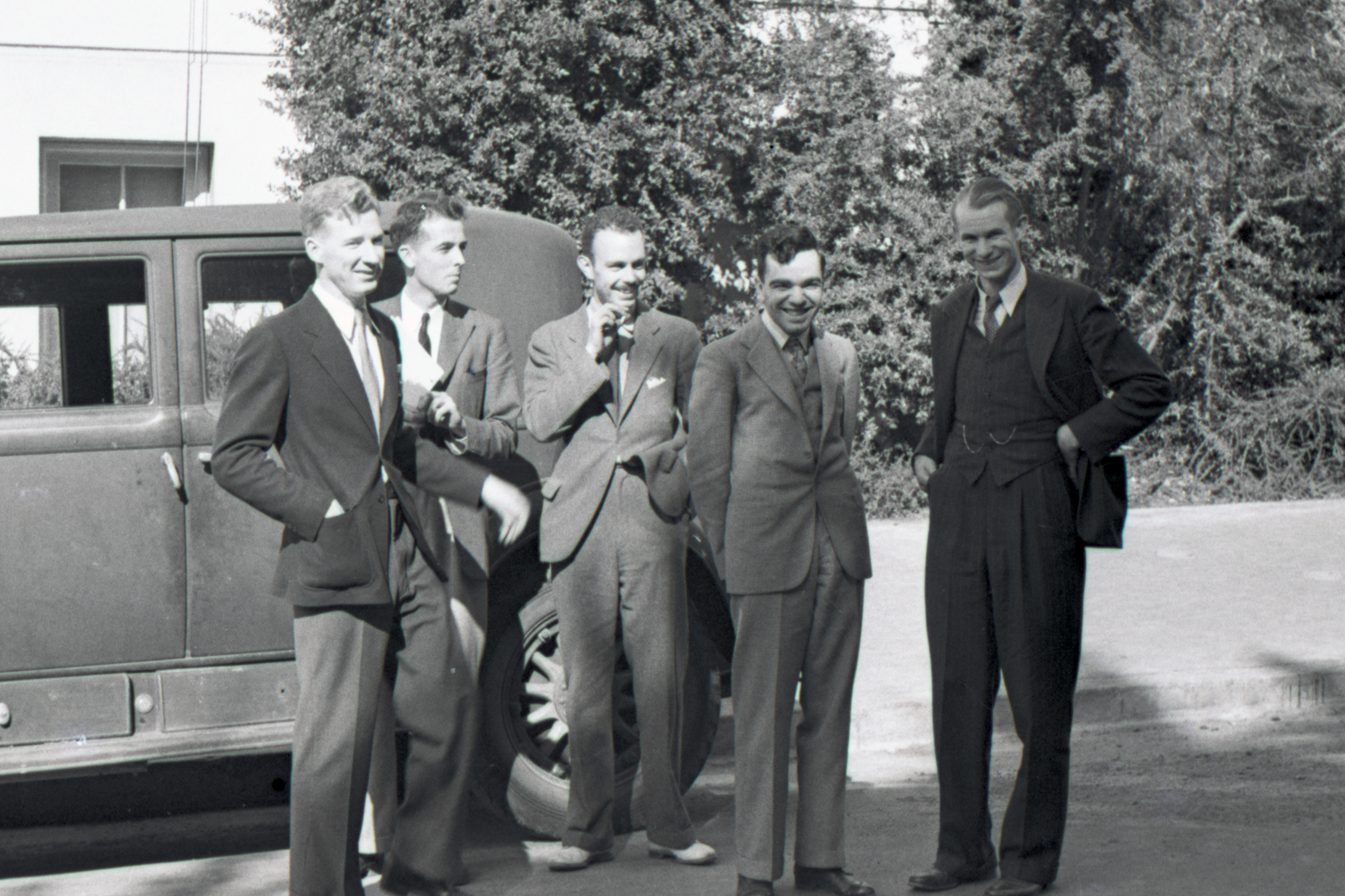 black and white photo of five men in coat and tie in front of vehicle and trees