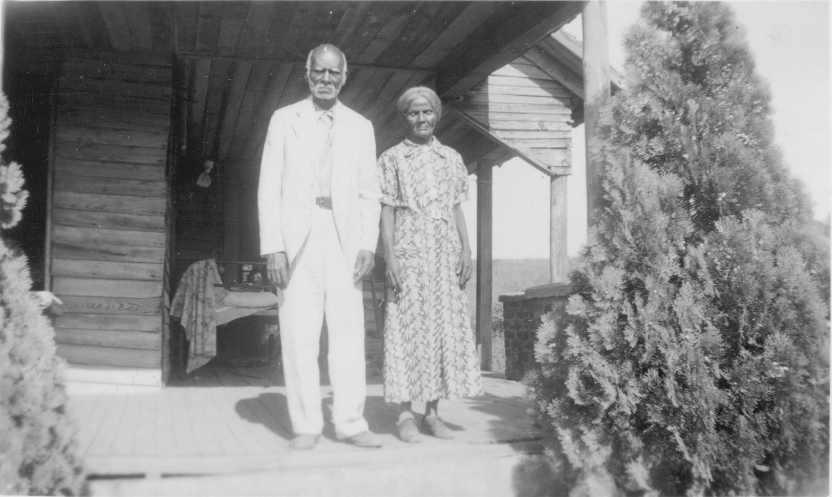 black and white photo of formally attired elderly Black couple standing in front of wooden house