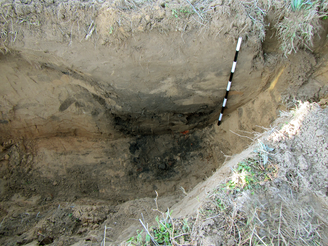 photo looking down into deep trench wall, at the base of which is a dark colored pit.