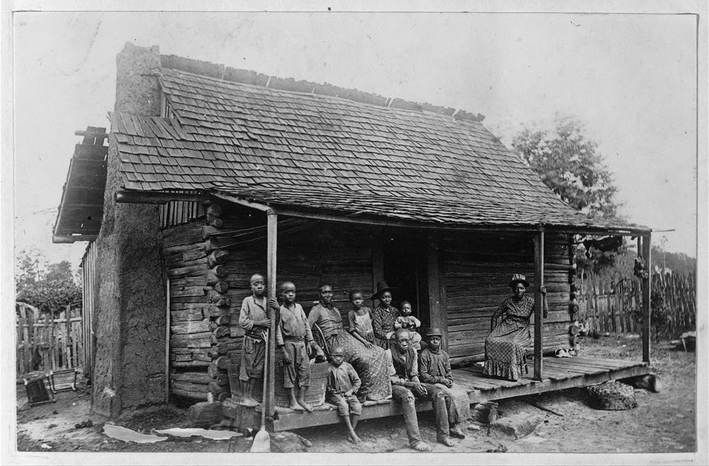 black and white photo of log cabin with large Black family sitting posed