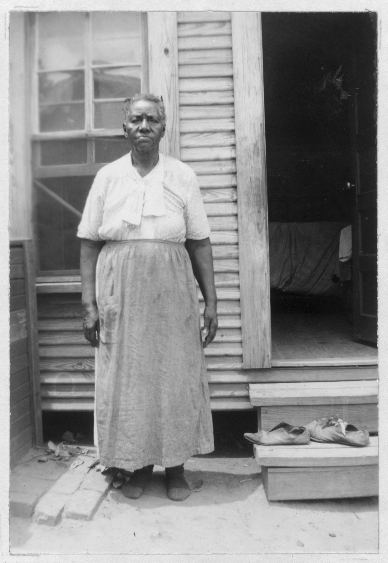 black and white photo of elderly Black woman standing in front of a raised wooden porch