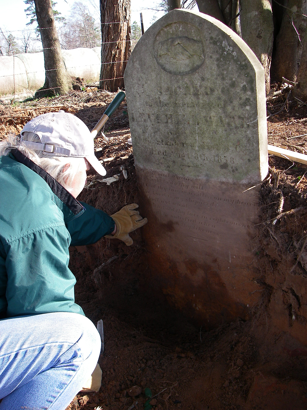 Photo of archeologist squating in excavation unit in front of headstone.