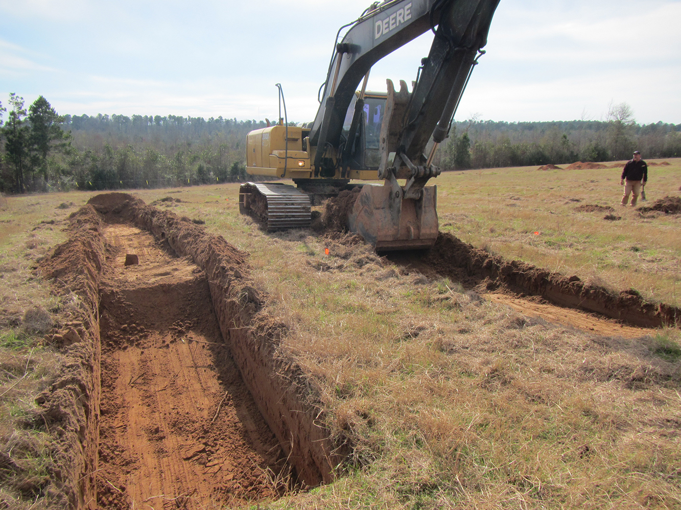work shot photo; on left is open trench excavated into red sandy soil; on right a large trackhoe is digging a second trench.